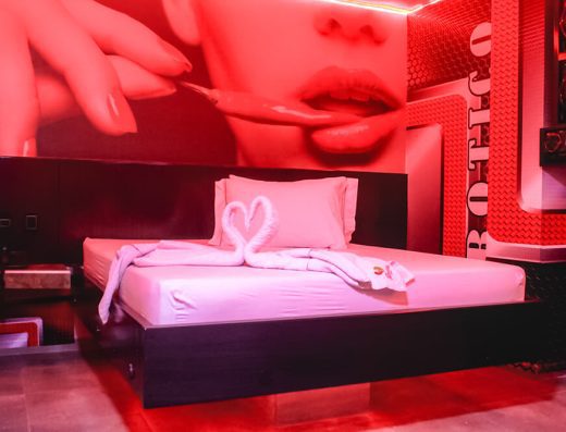 Habitación moderna de motel bañada en luz roja, decorada con arte mural de labios y grandes altavoces junto a una cama con lino blanco.